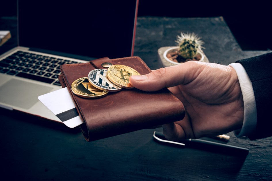 Homem segura carteira com bitcoins no topo, perto de mesa com computador