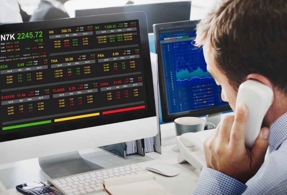 Homem fala ao telefone em frente a dois monitores com gráficos de investimentos