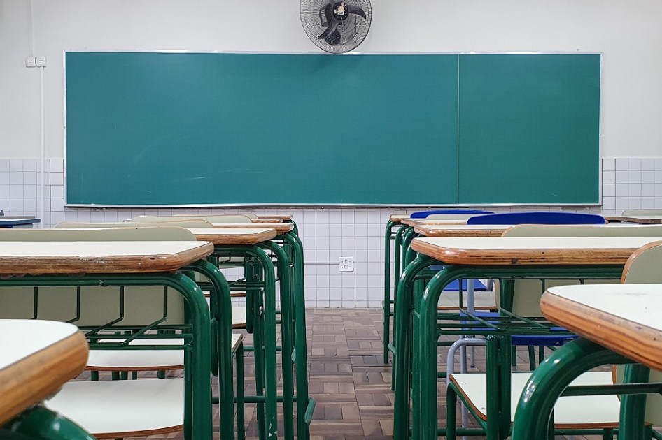 Vista de trás de uma sala de aula, com a lousa de frente para quem olha a imagem e um vão entre as carteiras, alinhadas lado a lado