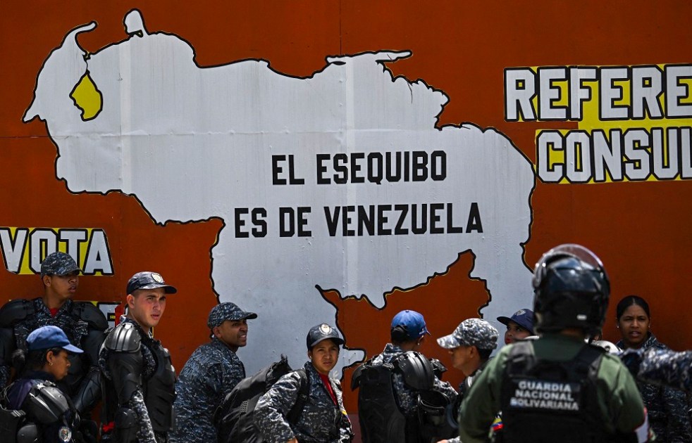 Integrantes da Polícia Nacional Bolivariana e da Guarda Nacional estão ao lado de um mural com o mapa da Venezuela e a frase "O Essequibo pertence à Venezuela" durante um protesto do Dia do Trabalhador em Caracas