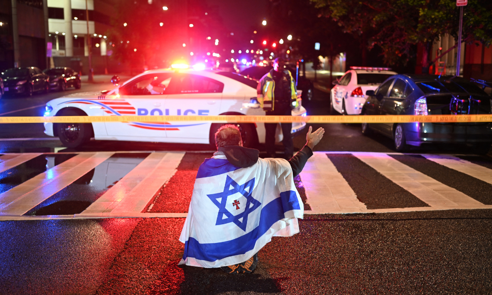 Um homem envolto na bandeira de Israel, carregando uma cruz com o nome "Jesus" no centro, gesticula enquanto policiais da Polícia Metropolitana isolam a área ao redor do Museu Judaico da Capital após um tiroteio que deixou duas pessoas mortas em Washington, DC, na madrugada de 22 de maio de 2025. Dois funcionários da embaixada de Israel foram mortos a tiros na noite de quarta-feira, 21 de maio, do lado de fora de um museu judaico em Washington, por um atirador que gritava "Palestina livre", segundo as autoridades. Autoridades americanas e diplomatas israelenses expressaram choque e indignação com os assassinatos.