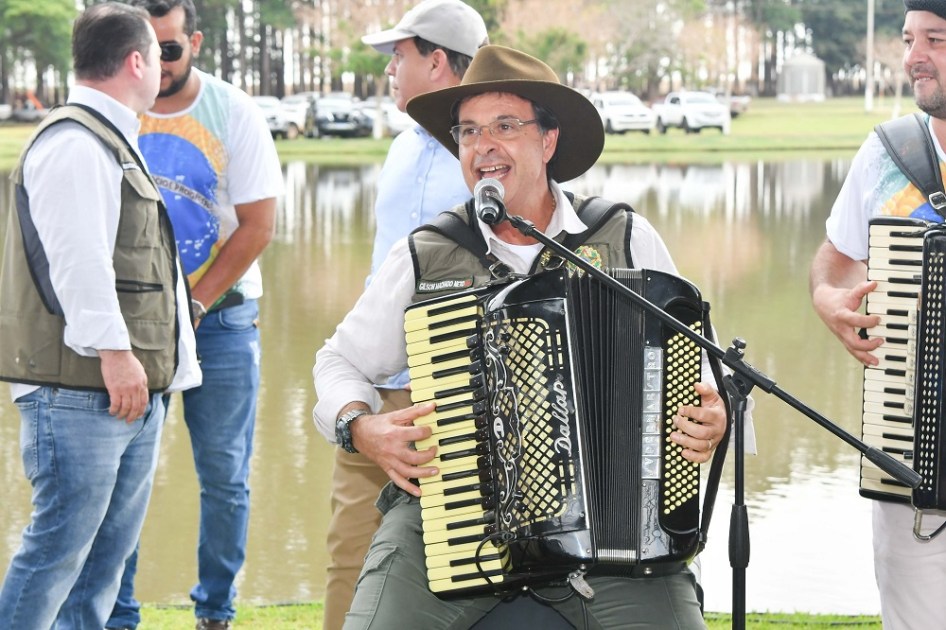 Sentado à frente de um microfone, usando chapéu, óculos,, colete e calça, o ministro Gilson Machado toca sanfona e canta a céu aberto, com outro sanfoneiro à sua esquerda; atrás dele, três pessoas conversam