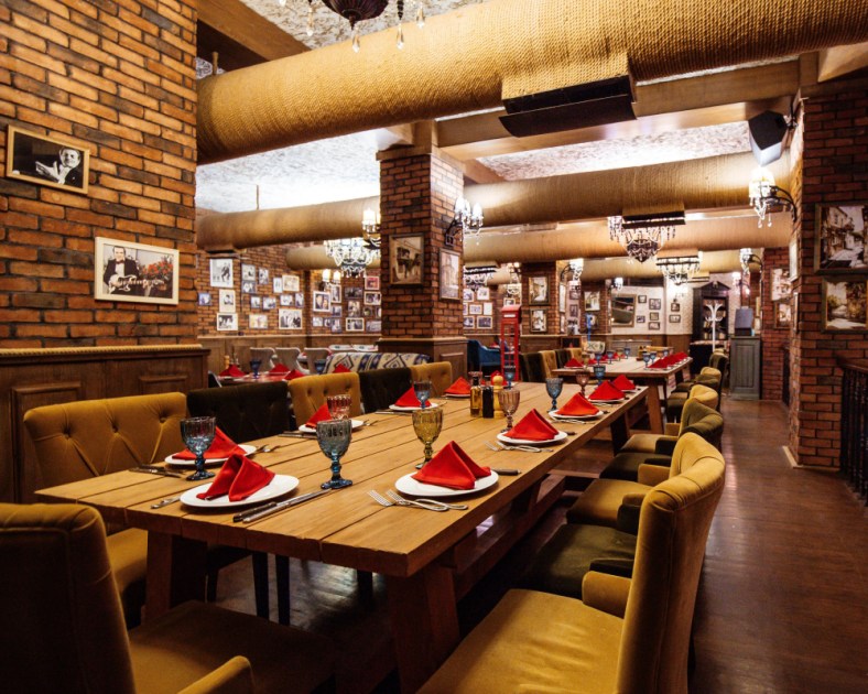 Uma sala de restaurante com paredes de tijolo vermelho, mesas de madeira e canos no teto