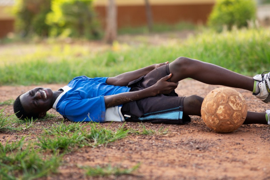 Jovem caído e com a mão no joelho em campo de futebol, ao lado da bola