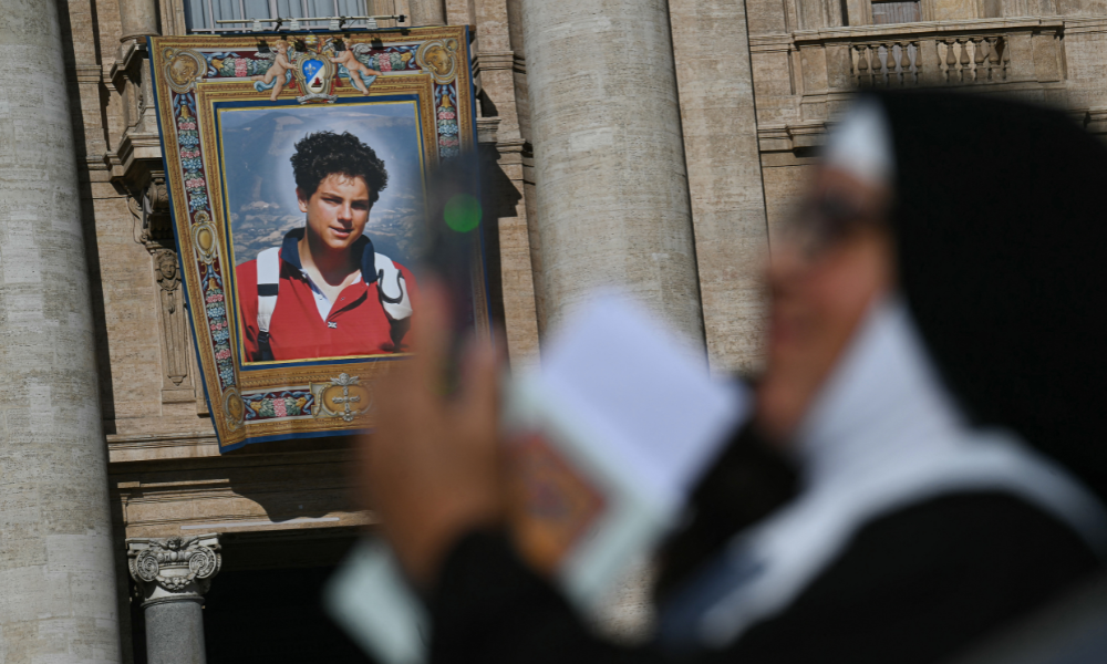 Uma freira verifica seu celular enquanto um retrato do falecido adolescente italiano Carlo Acutis é exibido na fachada da Basílica de São Pedro, ao fundo, antes da Santa Missa e da canonização do Beato Carlo Acutis e de Pier Giorgio Frassati na Praça de São Pedro, no Vaticano, em 7 de setembro de 2025. Um adolescente italiano apelidado de "Influenciador de Deus" por seus esforços para espalhar a fé católica online se tornará o primeiro santo milenar neste domingo, em uma canonização com a presença de milhares de peregrinos. O gênio da computação Carlo Acutis, que morreu de leucemia em 2006 aos 15 anos, será canonizado pelo Papa Leão XIV em uma cerimônia solene na Praça de São Pedro, no Vaticano. O italiano Pier Giorgio Frassati, um entusiasta do montanhismo que morreu em 1925 e era conhecido por seu compromisso social e espiritual, também será canonizado em 7 de setembro de 2025. (Foto de Filippo MONTEFORTE / AFP)
