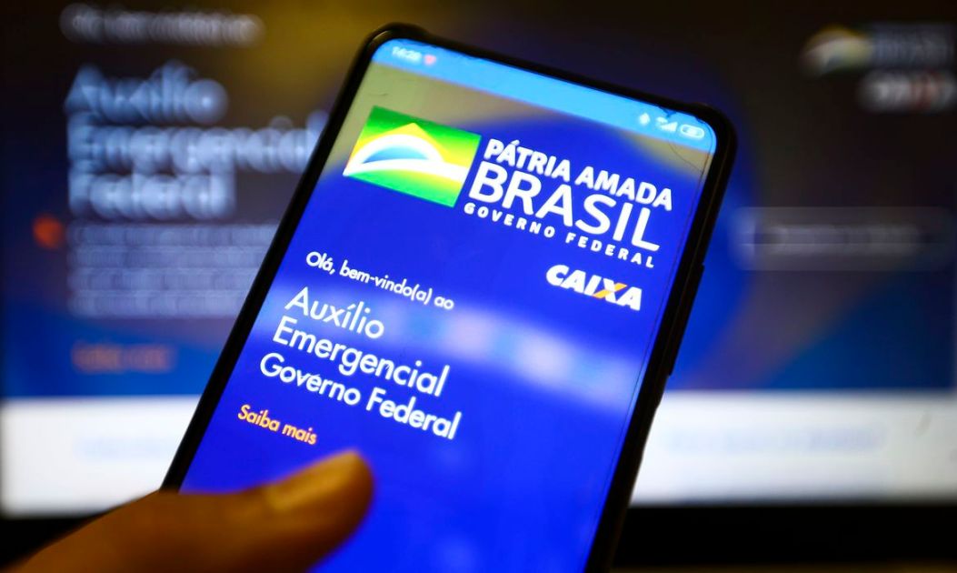 Mão segura smartphone com o aplicativo do auxílio emergencial