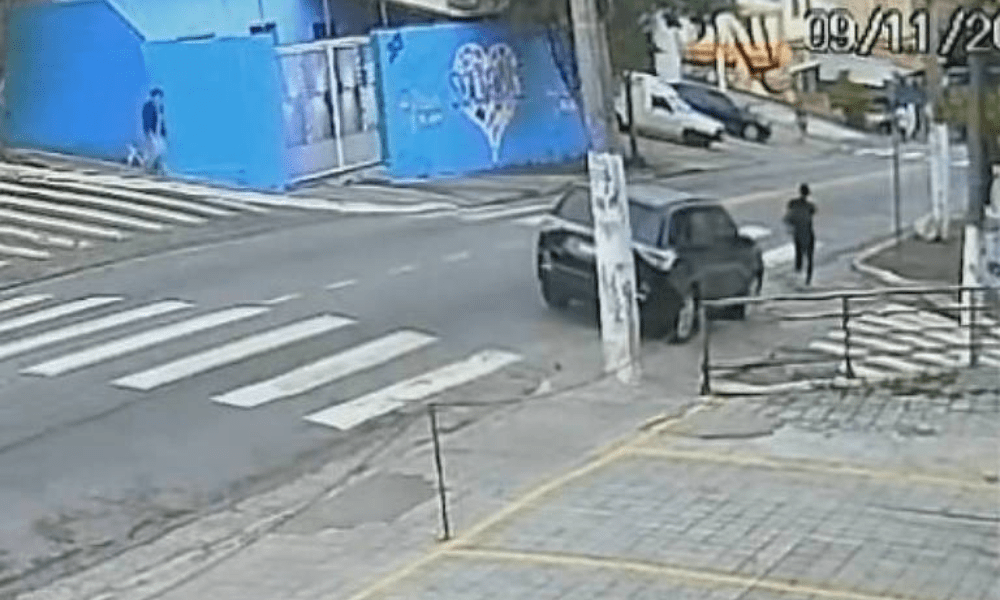 Homem está parado ao lado de carro preto estacionado próximo a esquina de rua com faixa de pedestres. Ambiente urbano com calçada e muro azul ao fundo. Momento em que mulher caminha pela calçada e veículo se aproxima segundos antes do atropelamento - Reprodução/TV globo