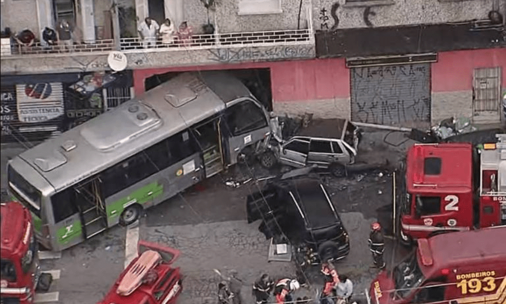 Ônibus colidido e tombado parcialmente na calçada, com dois carros amassados próximos. Equipes de bombeiros e veículos de emergência no local atendem a ocorrência. Pessoas observam da calçada e sacada de prédio próximo. Após colisão, micro-ônibus invadiu um comércio, na zona norte de São Paulo - Reprodução/TV Globo