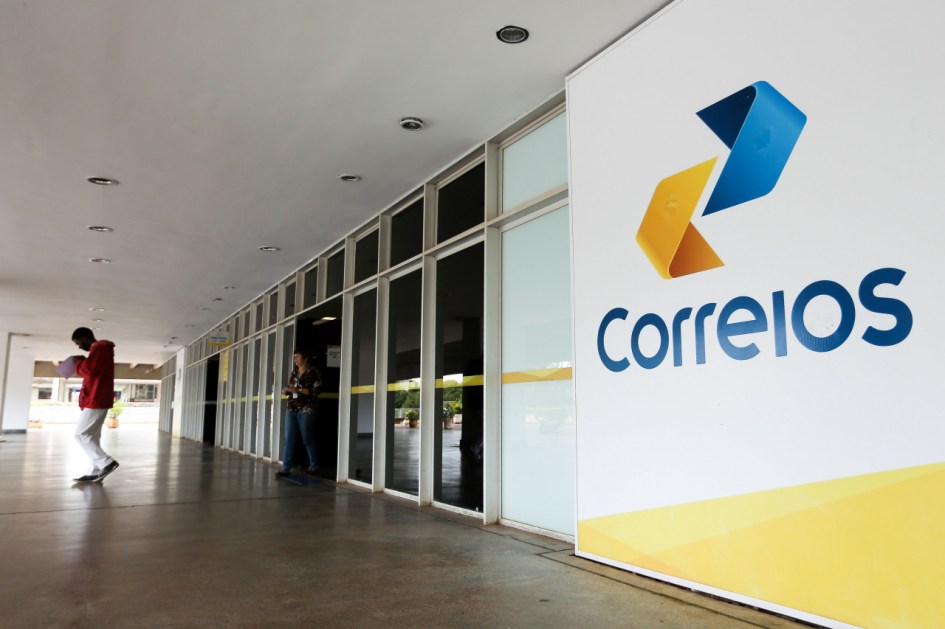 Fachada do Correios com o logo e uma parede branca com uma parte amarela. Tem uma porta de vidro e uma pessoa caminha
