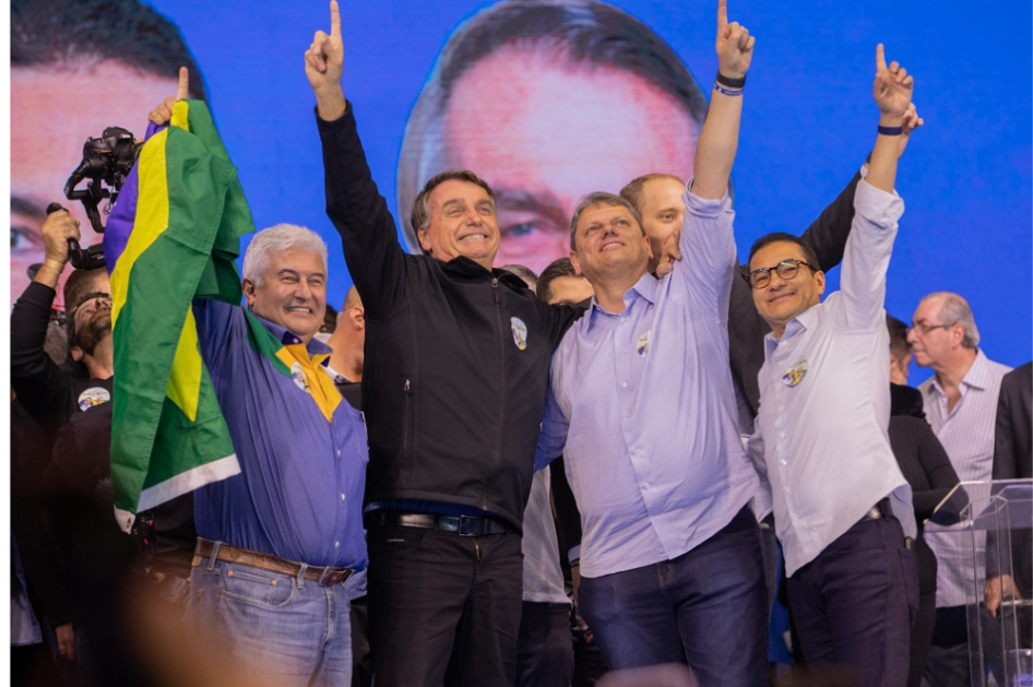 Marcos Pontes, segurando uma bandeira, Bolsonaro e Tarcísio, os três em pé, abraçados e apontando para o alto