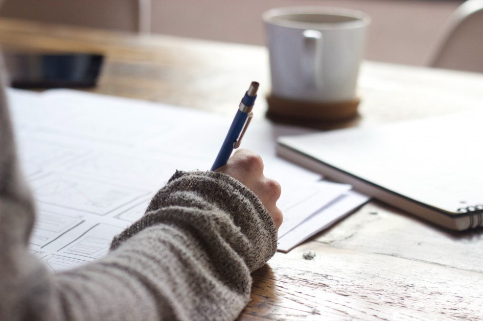 Mão anotando em um caderno com uma xícara de café atrás