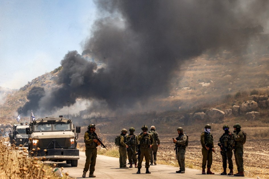 Forças de segurança israelenses montam guarda ao longo de uma estrada enquanto, ao fundo, fumaça surge de uma área supostamente incendiada por colonos israelenses na vila palestina de Idna, a oeste de Hebron, na Cisjordânia ocupada, em 5 de junho de 2026